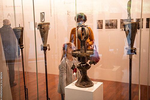 Au premier plan, quatre objets, dont trois bâtons en fer surmontés d'une forme d'entonoir. La photo est prise à travers la vitrine transparante et de l'autre côté, on voit une petite fille, le regard vers le haut, ainsi que deux adultes en arrière plan, de dos.