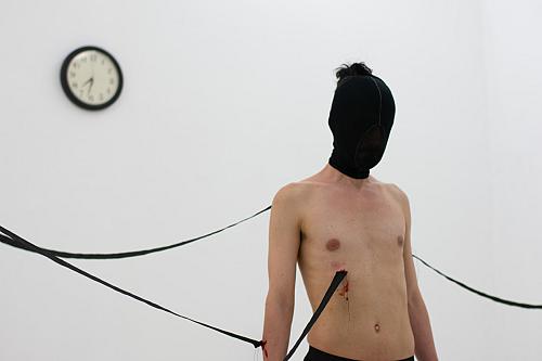 Une personne, la tête couverte d'un tissu noir, pose au milieu d'une salle blanche. De longs fils ou bandes de tissus semblent sortir de son torse.