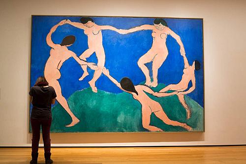 Devant à gauche, une femme se tient devant le tableau de Matisse, qui représente cinq femmes nues se tenant par la main, en cercle, sur de l'herbe verte et avec le ciel bleau au fond.